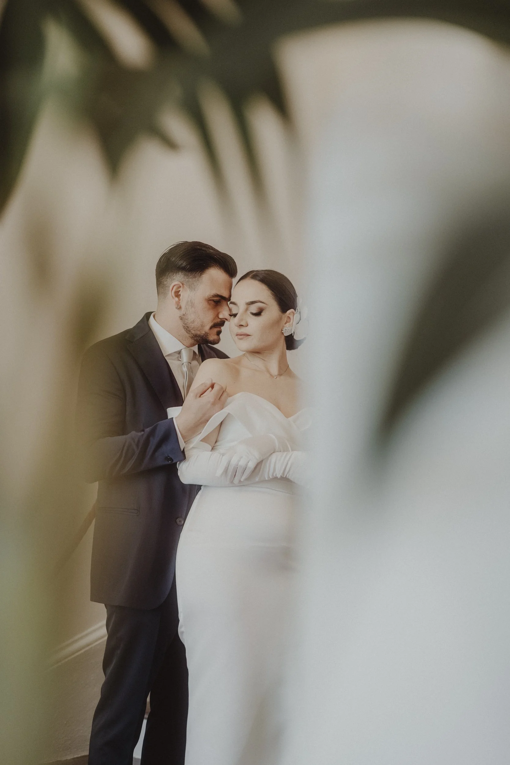 Sposi in abiti da matrimonio, visibilità limitata da foglie verdi in primo piano.