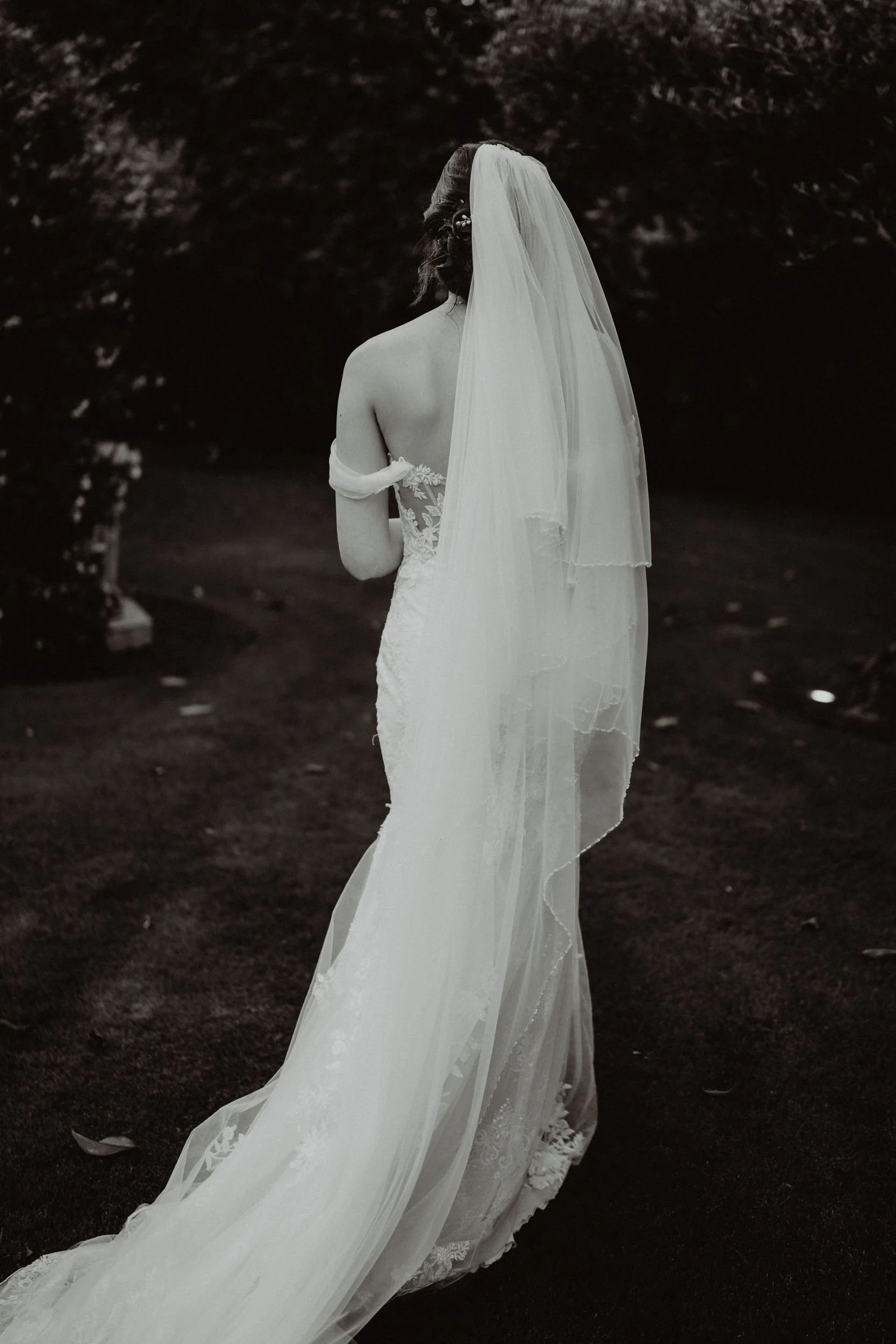 Donna in abito da sposa con lungale velo, vista di spalle, in un ambiente naturale.