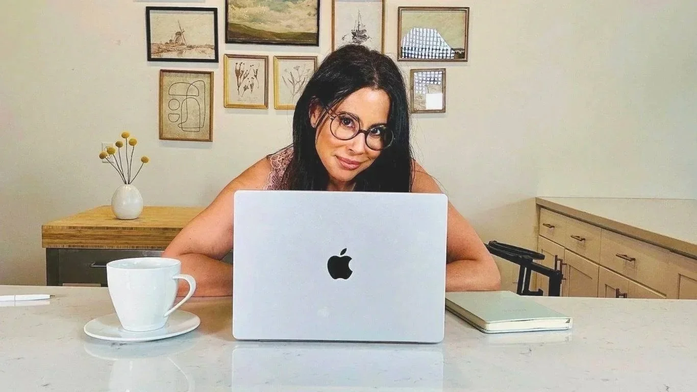 Melissa Maxwell, Content Strategist and Copywriter, with dark hair and glasses, sitting at a kitchen table with a laptop, coffee cup, and notebook, smiling at the camera with framed artwork on the wall behind her.