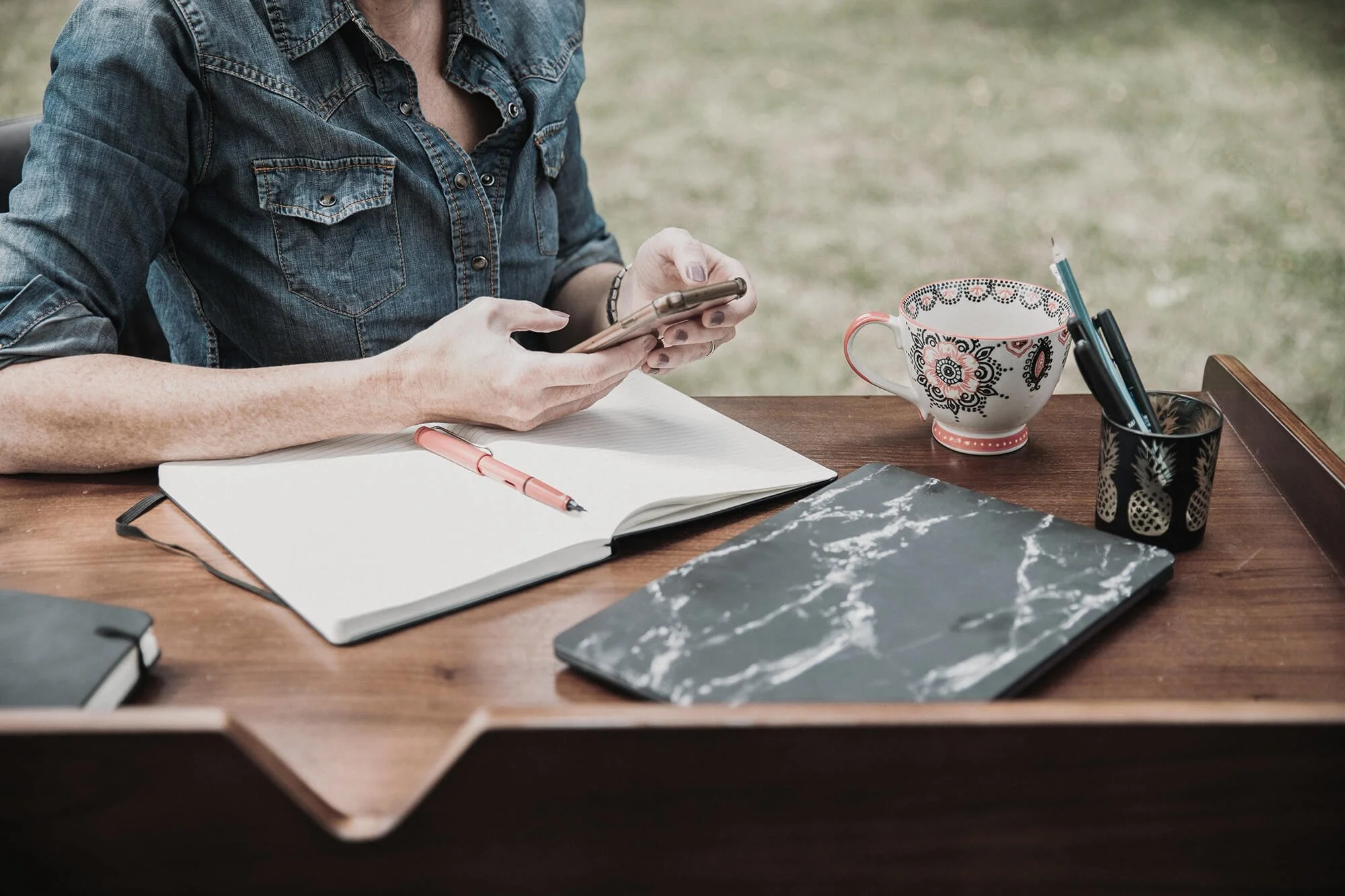 A person in a denim shirt sitting at a wooden desk, using a smartphone. The desk has an open notebook with a pink pen, a black notebook, a black container with pens, a patterned mug, and a marble laptop cover. The background appears to be outdoors with grass visible.