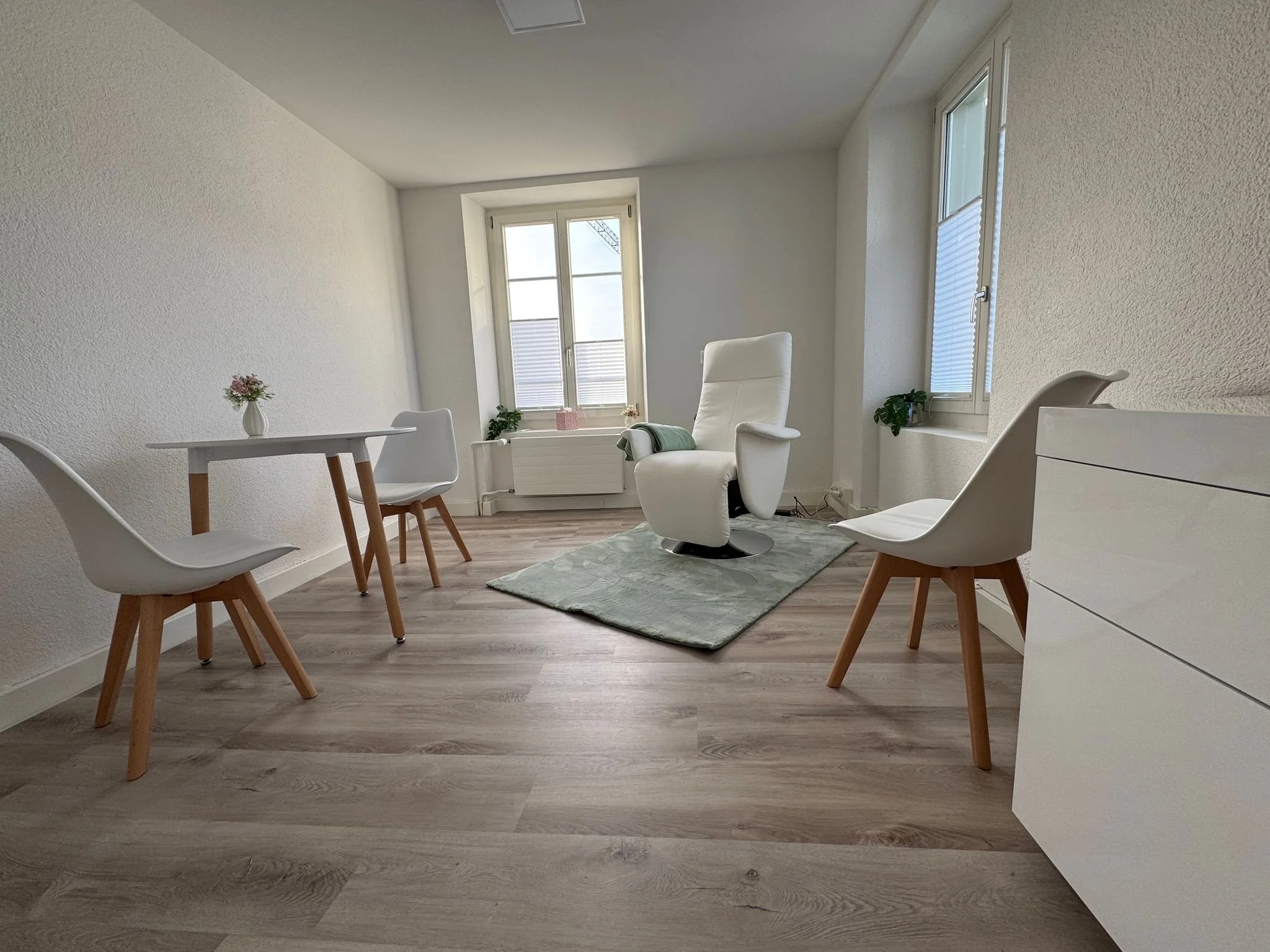 Minimalistischer, heller Raum mit weißen Möbeln, einem weißen gemütlichen Sessel, einem kleinen runden Tisch mit Vase und Blumen, Holzstuhl, Fenster mit Blick nach draußen, Holzfußboden, grüne Pflanzen