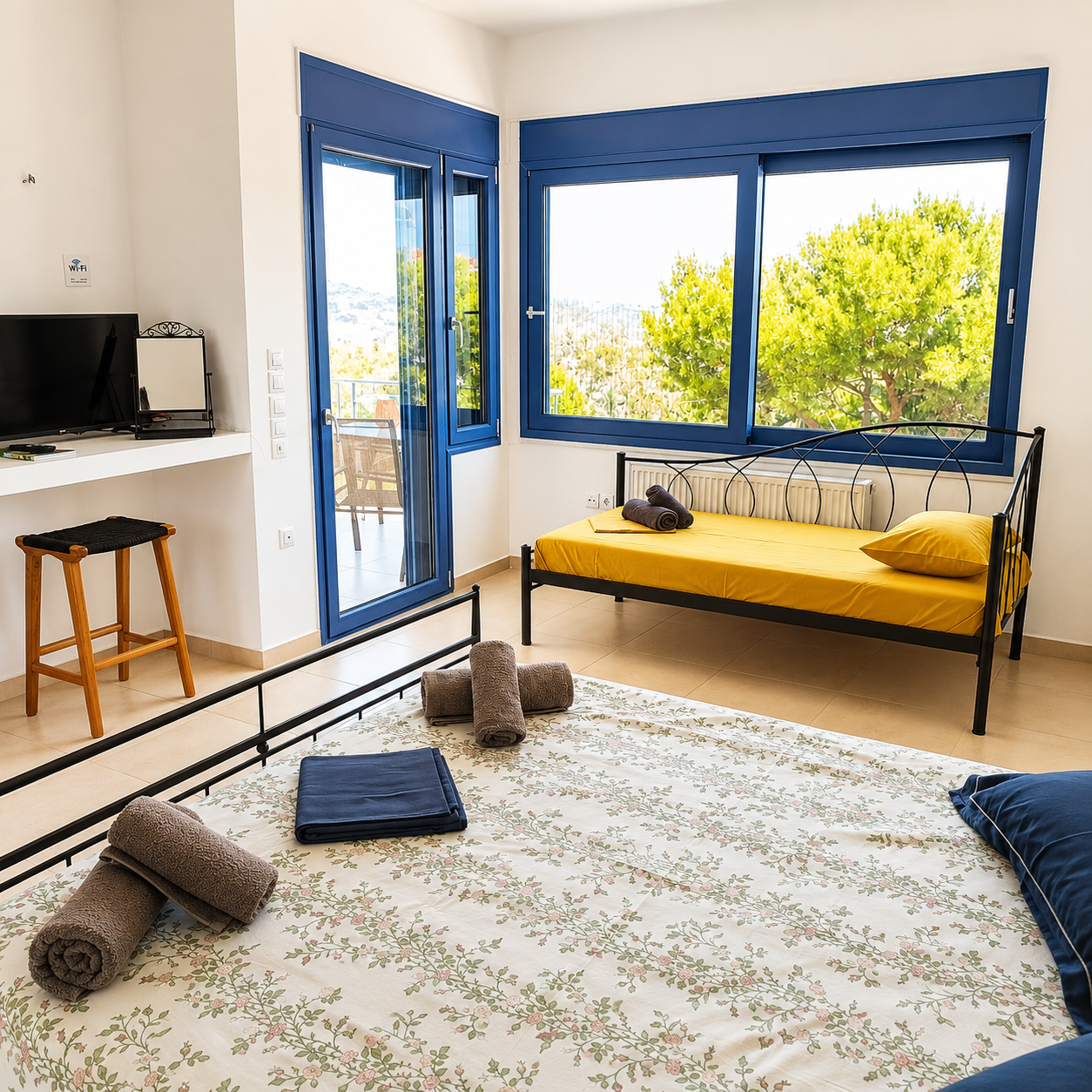 Bedroom with a bed covered in a blue and white dotted bedspread, pillows, and rolled towels. Small TV on a white shelf, a wooden stool, a wooden shelving unit, and glass door leading outside. Large windows with blue frames and a radiator below.