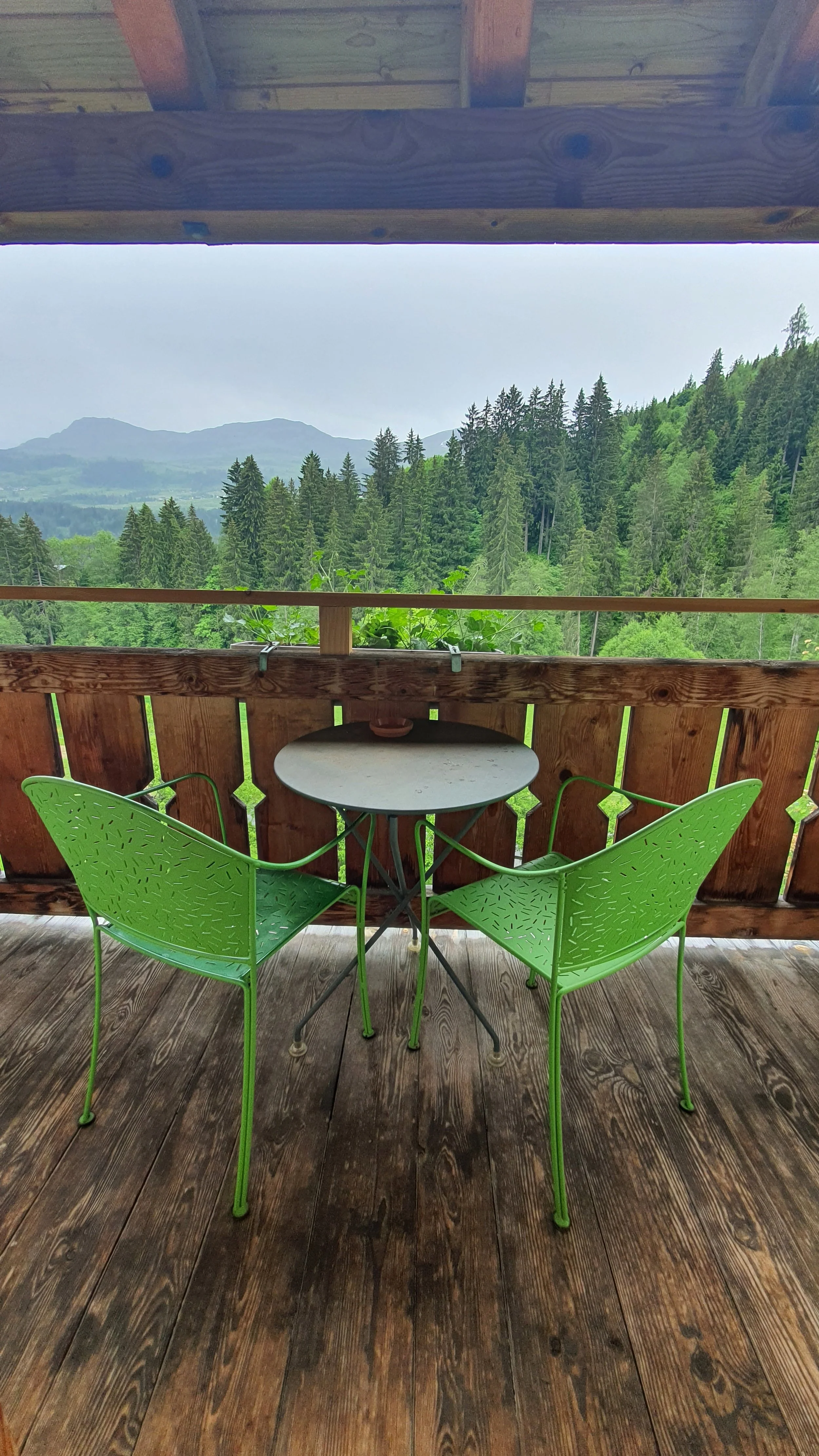 A small round table with two green chairs on a wooden porch, overlooking a lush green forest and distant mountains.