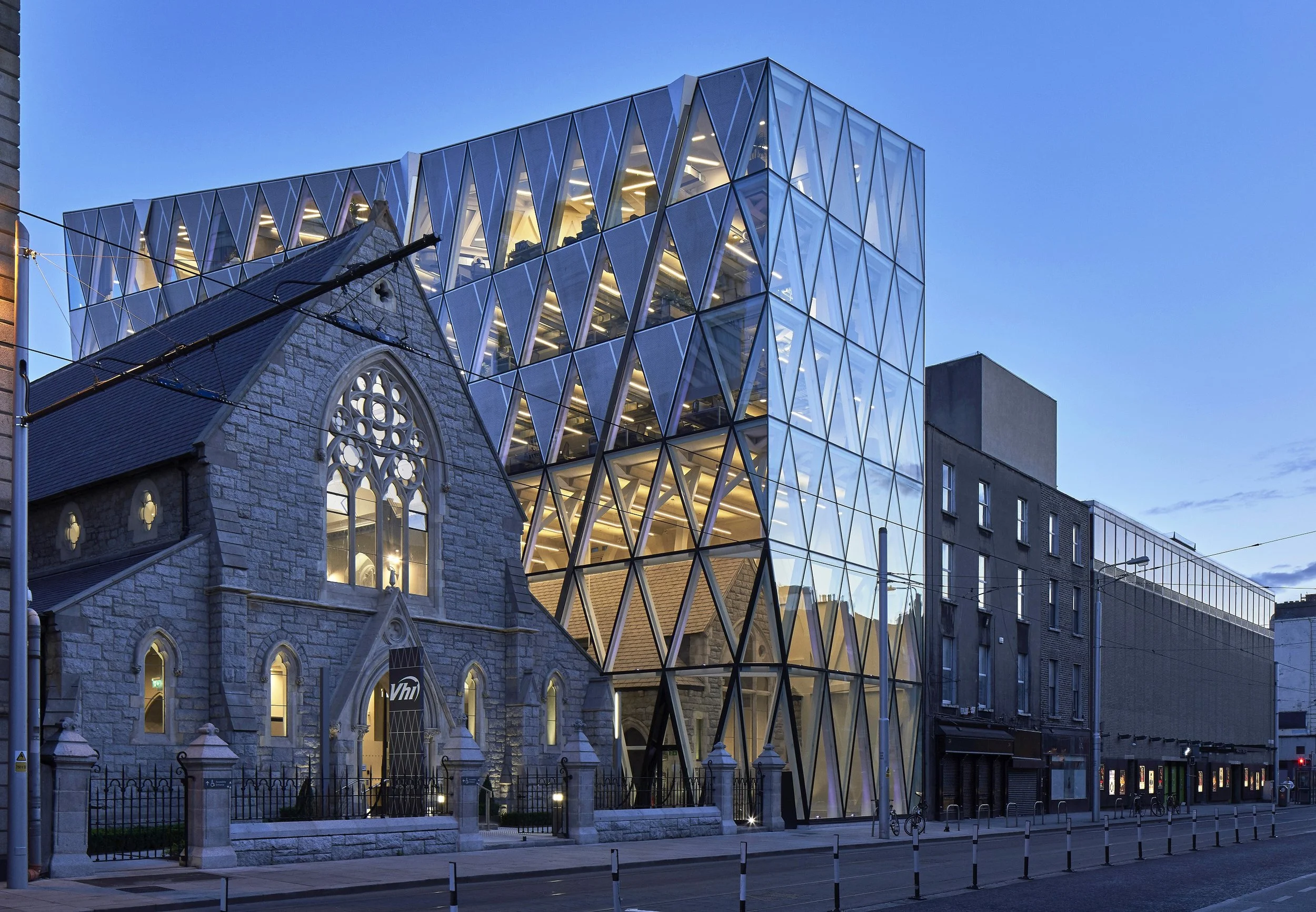 A historical stone church with arched windows and gothic architectural details is juxtaposed with a modern glass and steel building, creating a striking contrast of old and new architecture on a city street at dusk.