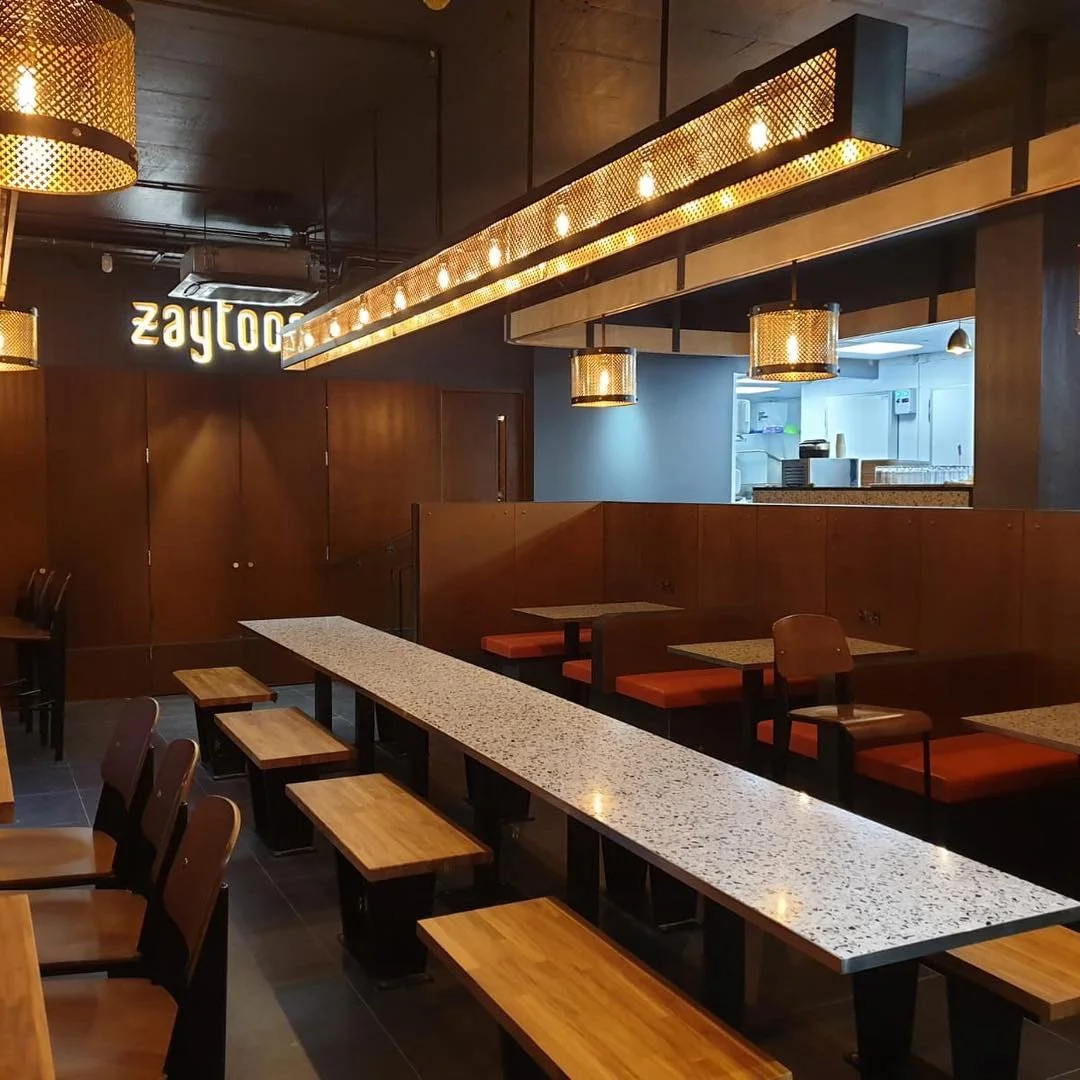 Interior of a modern restaurant with wooden tables and chairs, warm lighting, and a partially visible kitchen in the background.