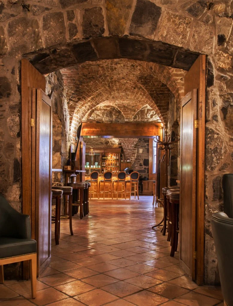 View through open wooden doors into a rustic bar with stone walls, arched ceiling, wooden bar counter, and leather chairs.