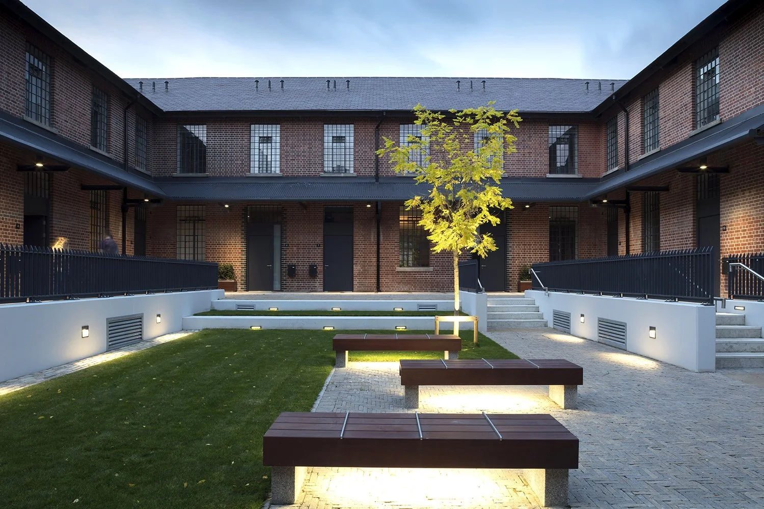 Modern apartment courtyard at dusk with benches, a small tree, and lighting