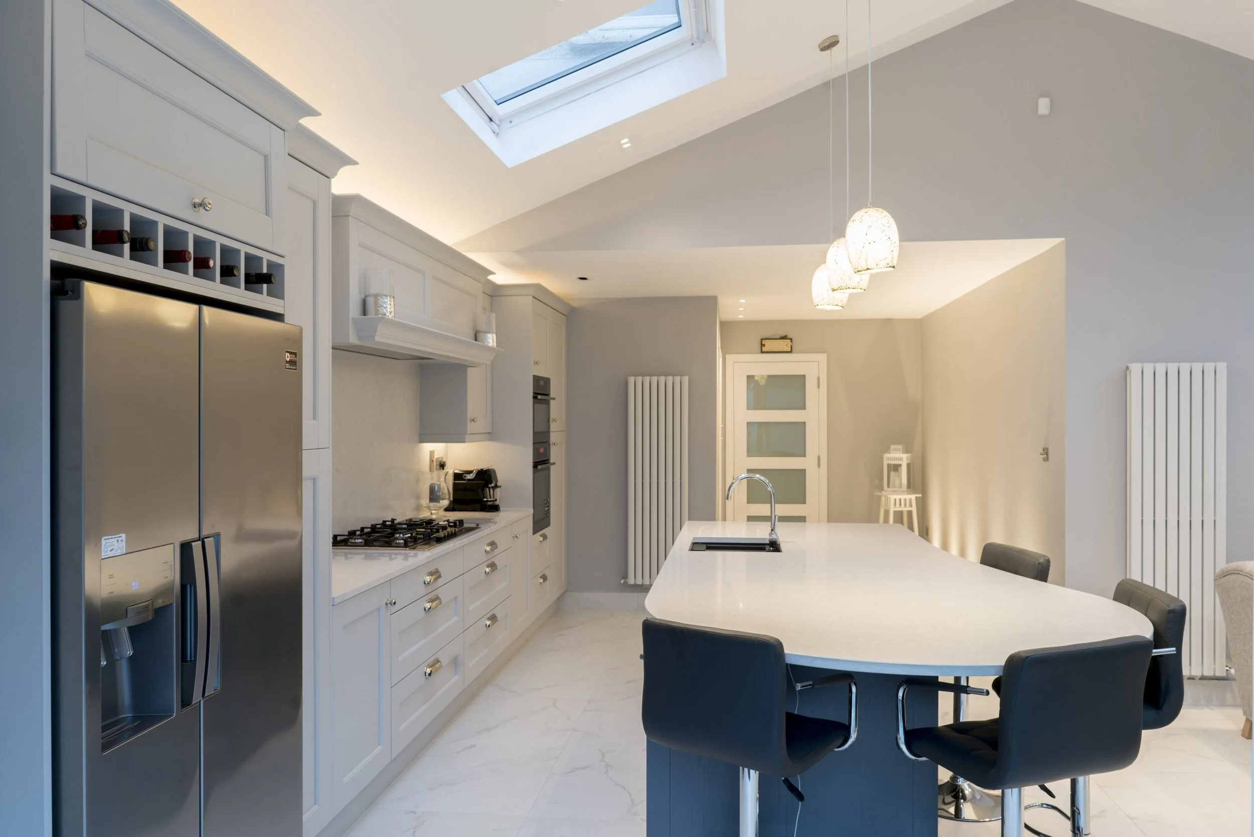 Modern kitchen with white cabinetry, stainless steel refrigerator, gas stove, built-in oven, and a large island with a white countertop and black chairs. Skylight and pendant lights illuminate the space.