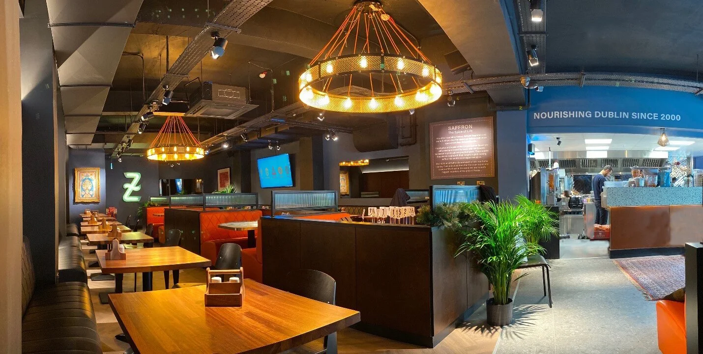 Interior of a modern restaurant with wooden tables, black chairs, and decorative lighting fixtures. There are potted plants and wall art, with a visible kitchen area in the background.