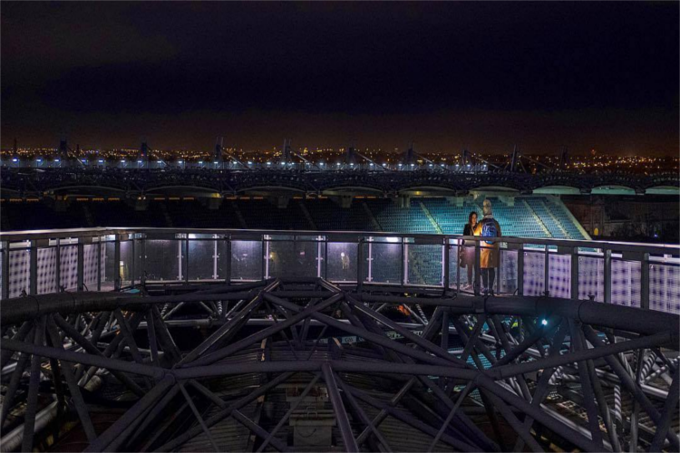 Nighttime view from an elevated observation deck or platform overlooking a cityscape with illuminated buildings and structures in the distance. Two people are standing near the railing on the right side, looking out at the city, with a city skyline a