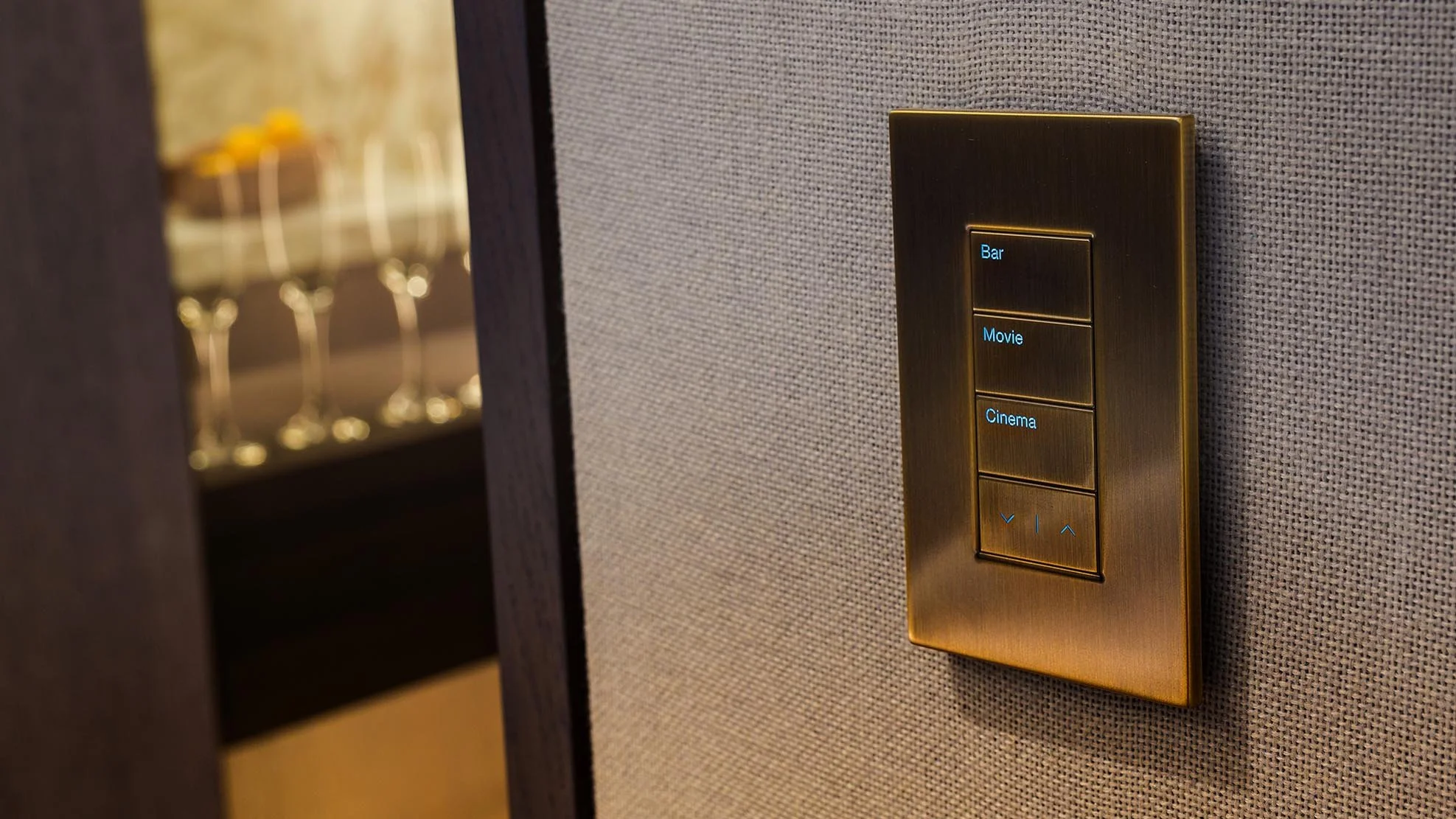 Close-up of a modern control panel with gold finish on a textured wall, featuring buttons labeled Bar, Movie, and Cinema, with arrow and volume controls beneath.