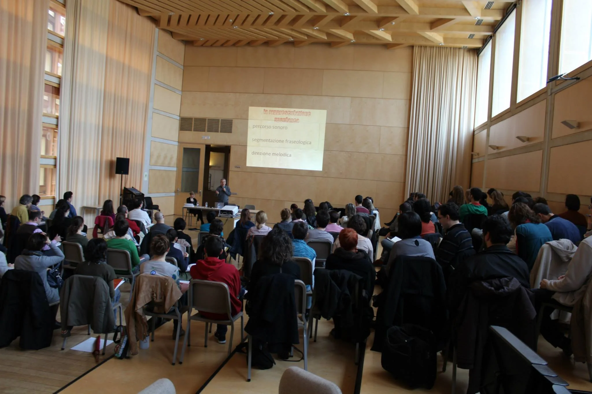 Conferenza con numeroso pubblico in una sala, presentatore davanti a uno schermo con slide in italiano - Alberto Odone.