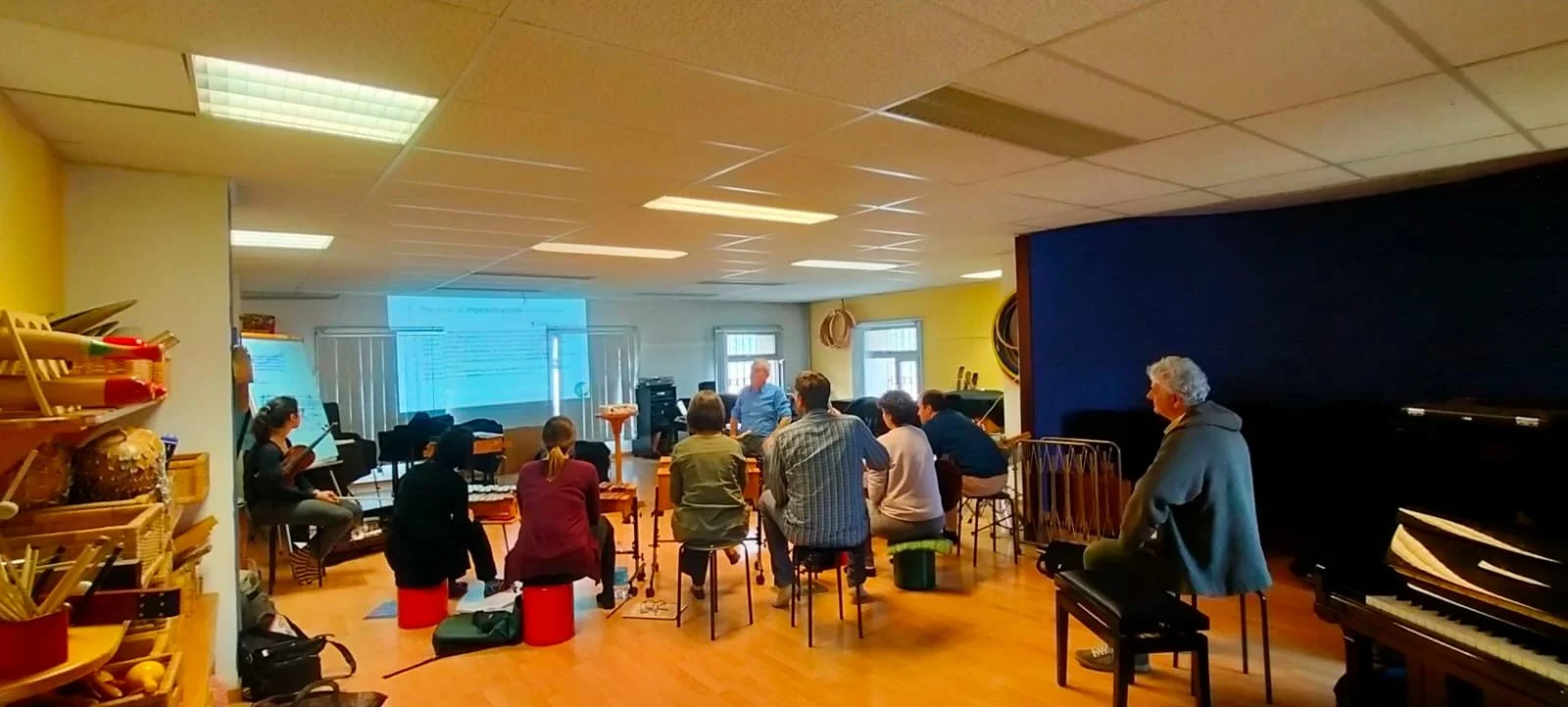 Un gruppo di persone sedute in cerchio in una stanza, in un corso o lezione di musica di Alberto Odone, con strumenti musicali e un pianoforte presenti.