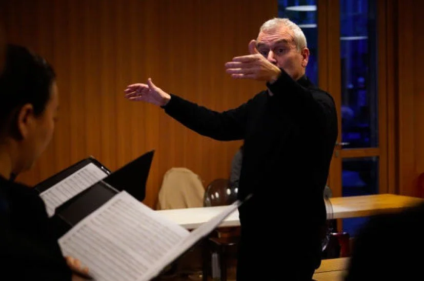 Alberto odone dirige un'orchestra in una sala di pratica o di registrazione. L'uomo ha i capelli grigi e indossa un abito nero, mentre sul tavolo ci sono alcuni strumenti e fogli di musica.