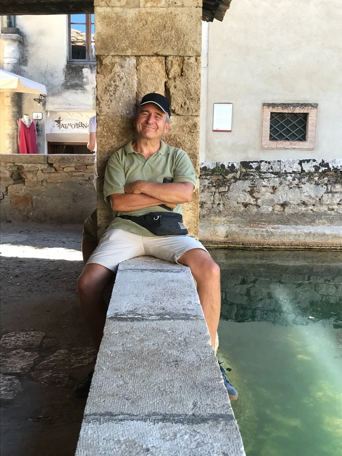 Uomo seduto con le braccia incrociate su un muretto di pietra vicino a un canale d'acqua, in un paesaggio urbano storico