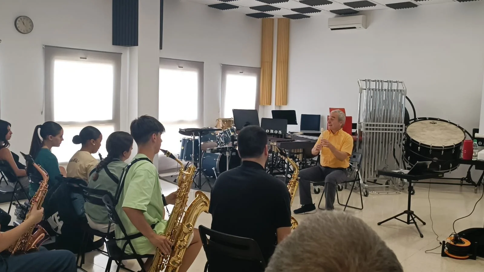 Gruppo di studenti con strumenti musicali in classe, ascoltando Alberto Odone, con strumenti come sax, batteria, tamburi e altri in sala prove.