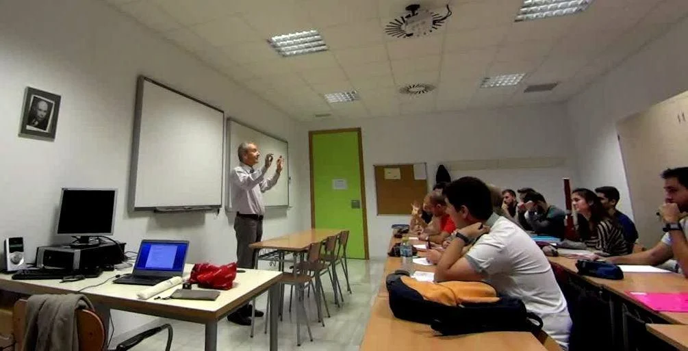 Classe di studenti in un'aula con Alberto Odone, con un insegnante davanti a una lavagna bianca, mentre alcuni studenti ascoltano e prendono appunti.