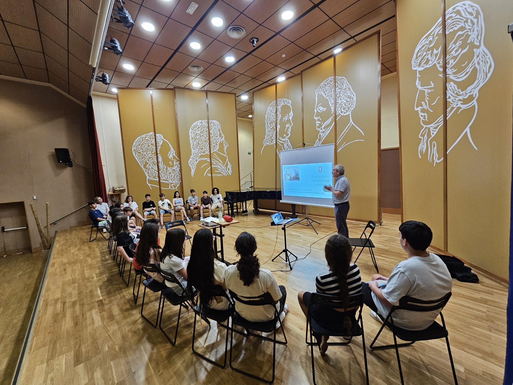 Alberto Odone presenta una lezione a un gruppo di studenti seduti in cerchio in una sala di scuola.