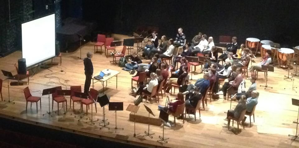 Un'orchestra si prepara per una prova o un concerto in un teatro, con vari strumenti e un pubblico di ascoltatori