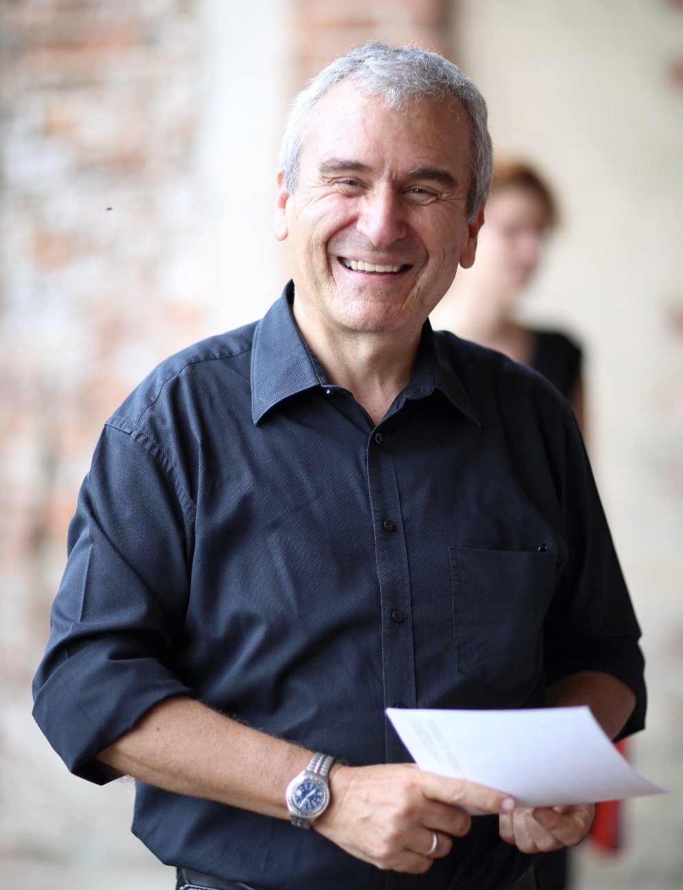 Uomo sorridente con camicia nera che tiene dei fogli, sfondo sfocato di pareti in pietra e altra persona sfocata sullo sfondo.