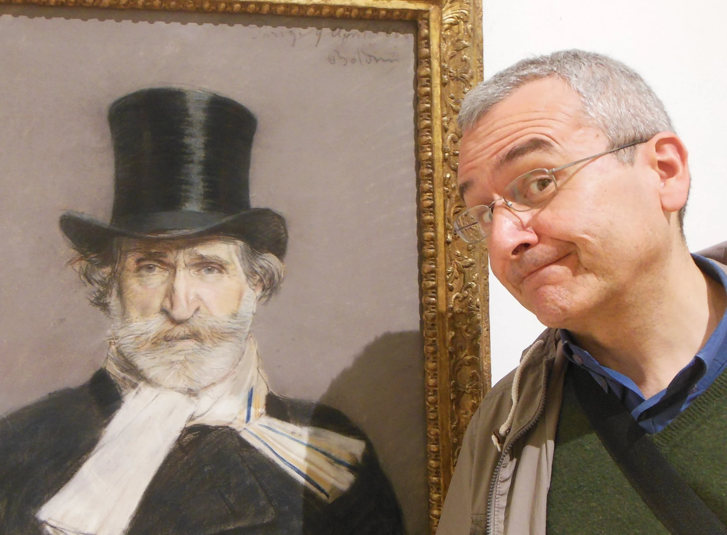 Un uomo con occhiali posa accanto a un ritratto di un uomo con cappello a cilindro, barba bianca e giacca scura.