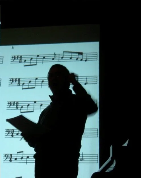 Silhouette di una persona che si trova davanti a una lavagna con partitura musicale proiettata, mentre tiene un foglio in mano e si sistemi i capelli.
