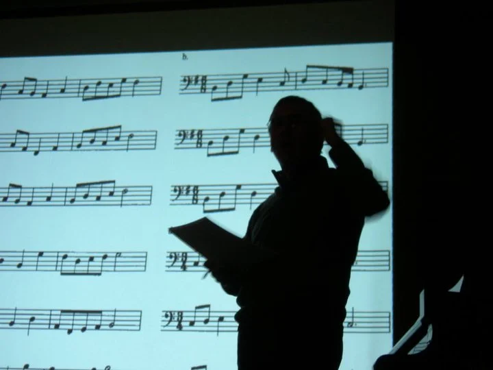 Figura di Albetrto Odone che tiene un foglio di carta davanti a uno schermo che mostra una partitura musicale.