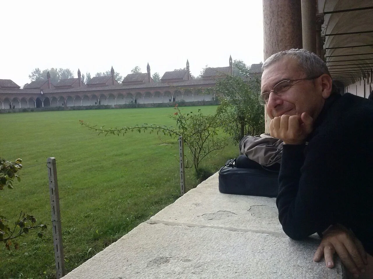 Un uomo con occhiali seduto su un muretto di pietra all'aperto, con un cortile verde e un edificio storico con archi sullo sfondo