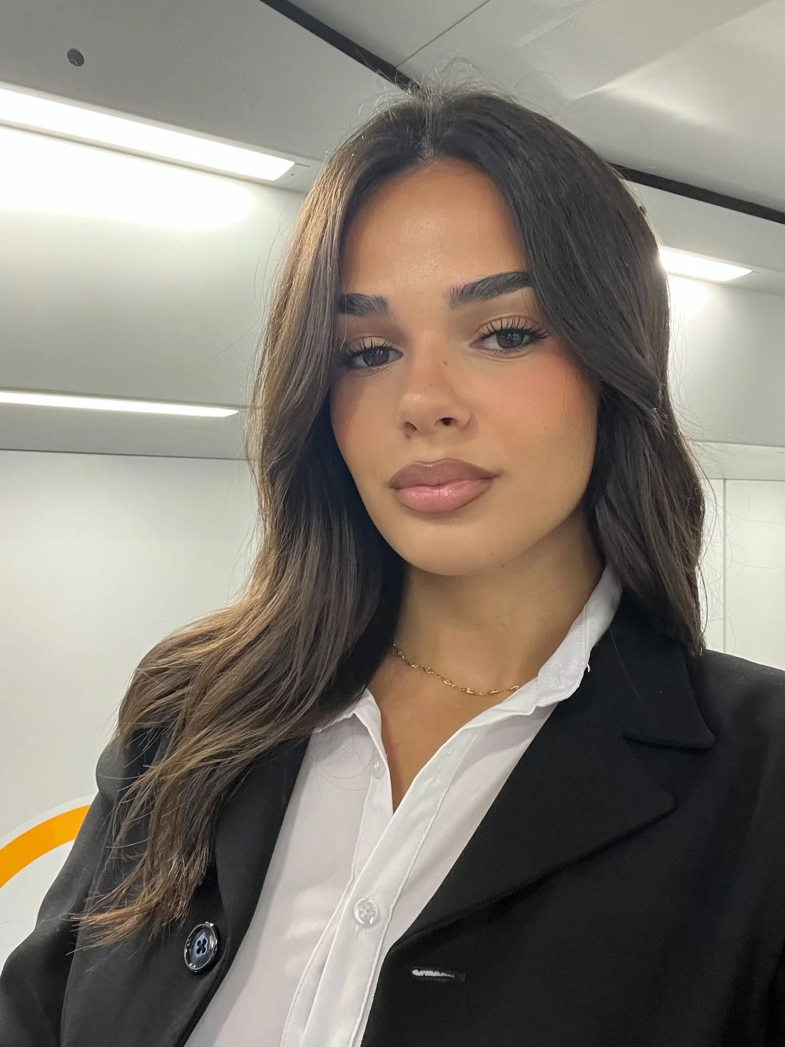 A woman with long dark wavy hair, wearing a black blazer over a white shirt, standing indoors with a neutral expression.