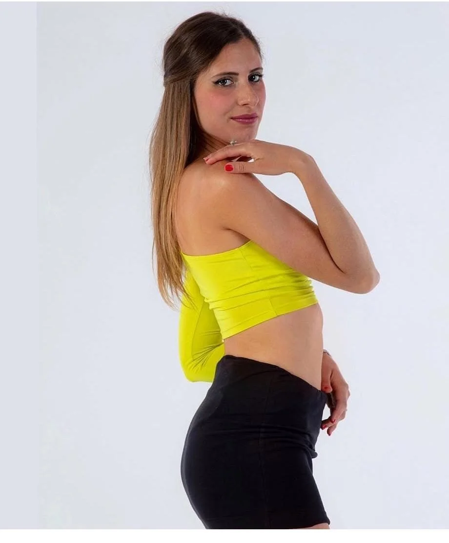 A woman with long brown hair wearing a yellow strapless top and black fitted shorts, standing sideways against a plain white background. She is touching her shoulder with one hand and placing the other hand near her hip, looking at the camera.