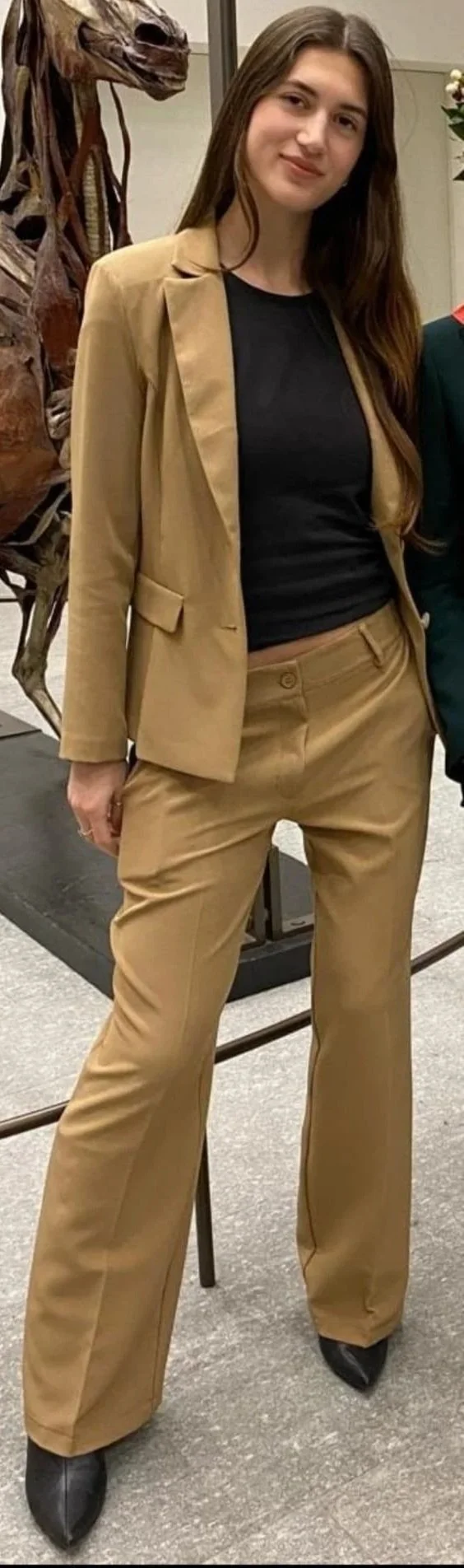 A young woman with long brown hair wearing a black top and tan suit, standing indoors near a dinosaur skeleton.