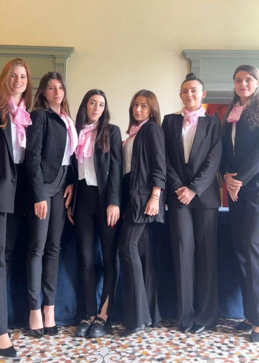 Group of six women standing side by side, dressed in black suits with pink scarves, indoors against a yellow wall with moldings.