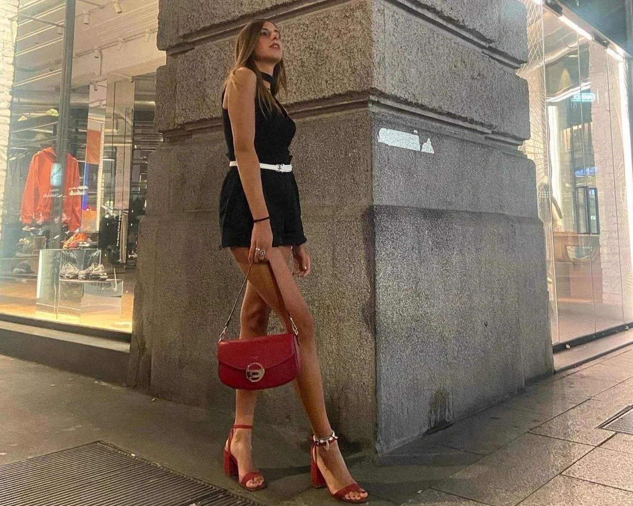 A woman standing on a city sidewalk at night, leaning against a stone building corner, wearing a black sleeveless top, black shorts with a white belt, red high heels, and carrying a red purse.