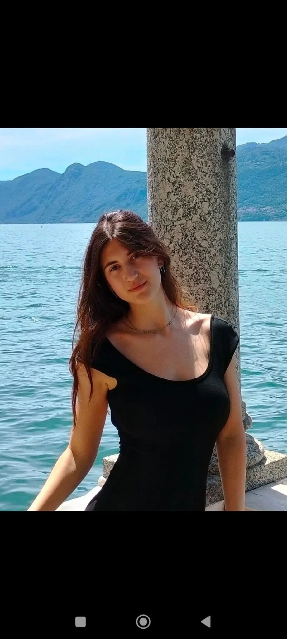 A young woman with long brown hair wearing a black sleeveless top, jewelry, and earrings, leaning against a stone pillar by the water with mountains in the background.
