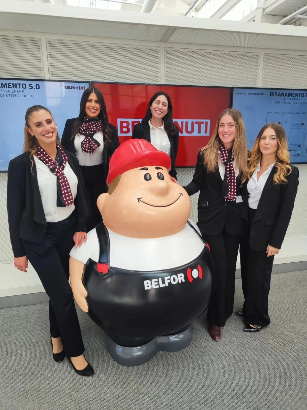Five women in business attire posing with a large cartoon statue of a construction worker wearing a red hard hat, against a backdrop of digital screens at an indoor event.