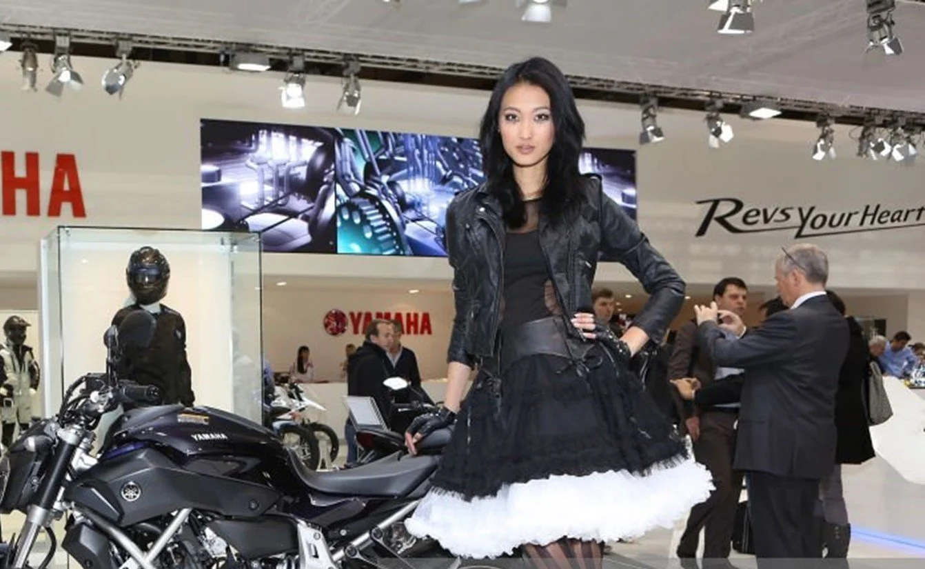 A woman with black hair in a black leather jacket and black skirt standing next to a black Yamaha motorcycle at a trade show, with people in the background and Yamaha branding visible.