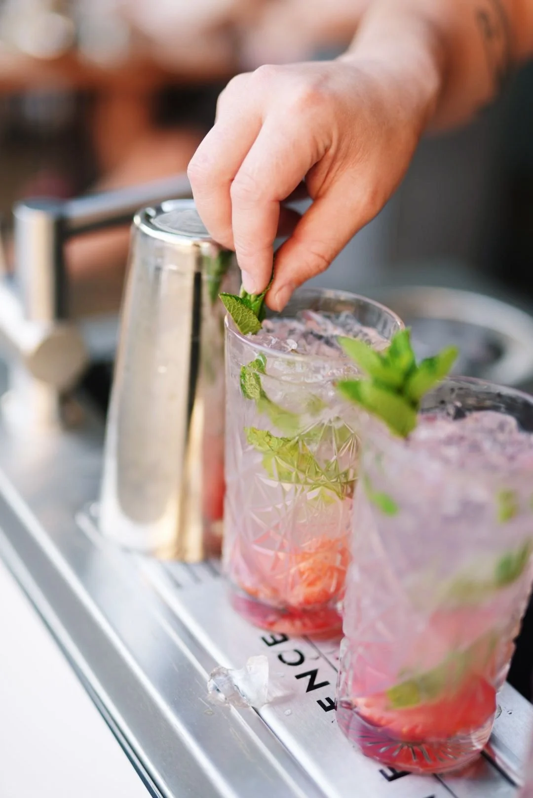 A person's hand adding a mint sprig to a tall glass of pink cocktail with ice and mint leaves, on a metal tray.