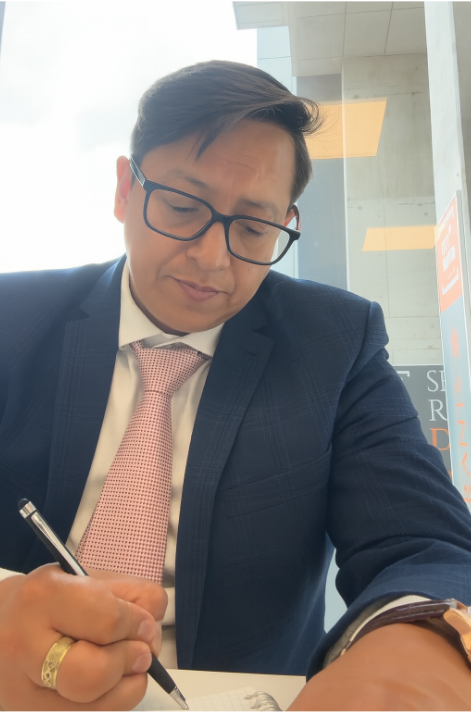 A man in a business suit and pink tie signing a document at a table in an indoor setting.