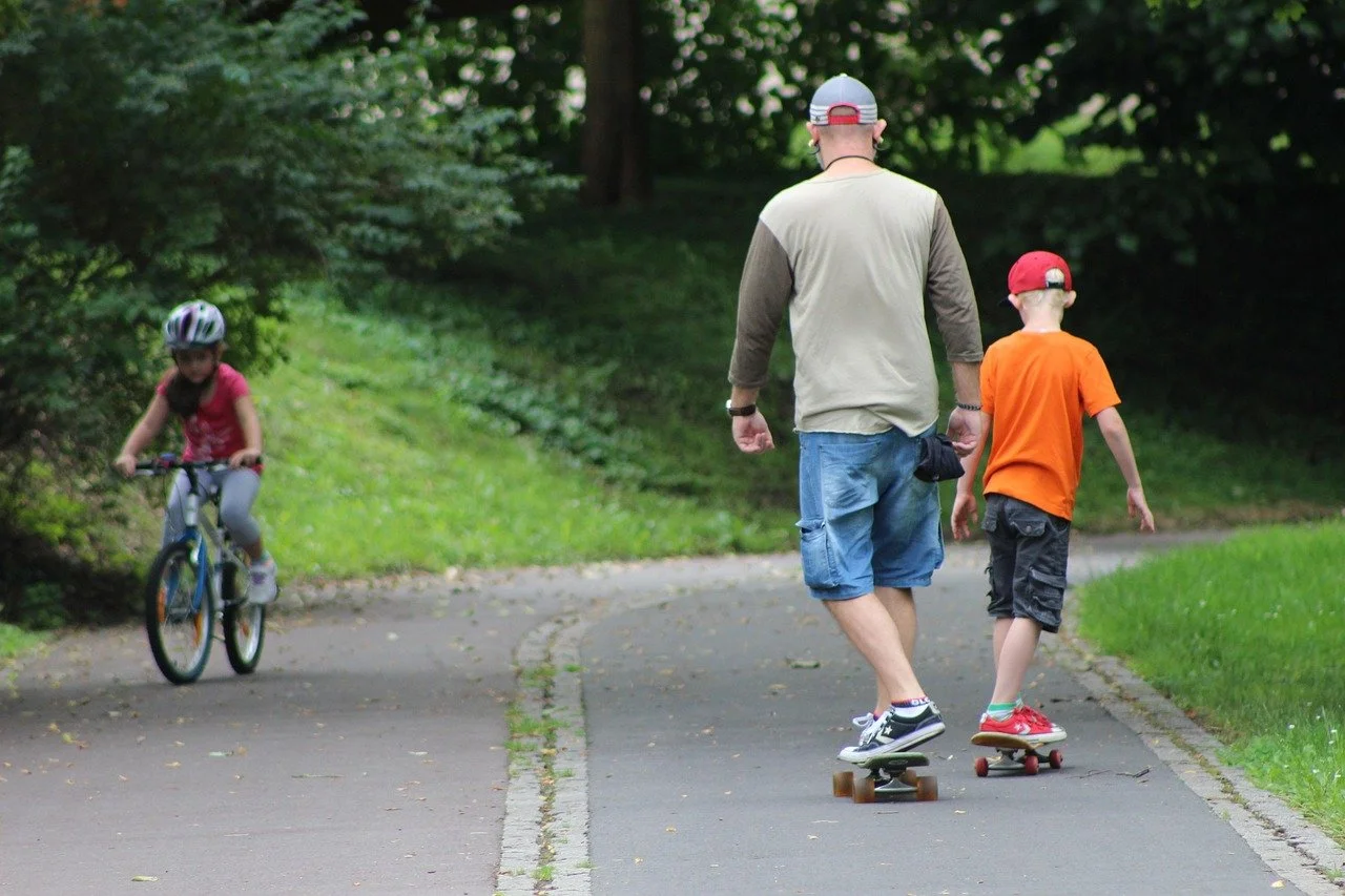 Un homme et un garçon font du skateboard sur un chemin dans un parc, pendant qu'une fille en vélo passe à proximité.