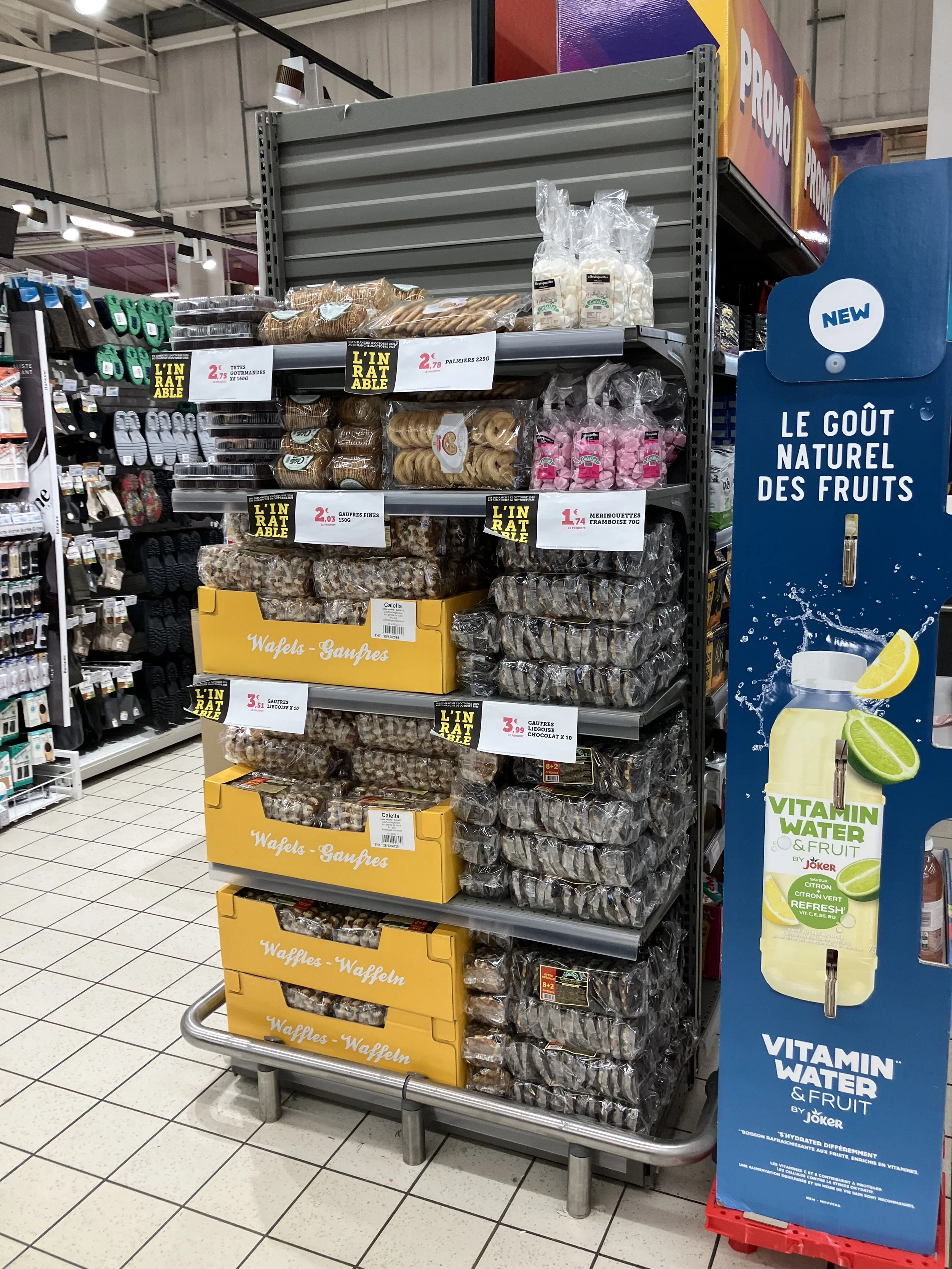 Présentoir de biscuits et de gaufres dans un supermarché, avec des emballages colorés et des prix indiqués. À droite, une affiche promotionnelle pour des boissons à base de fruits et de vitamine, avec une image de bouteille citron-lime.
