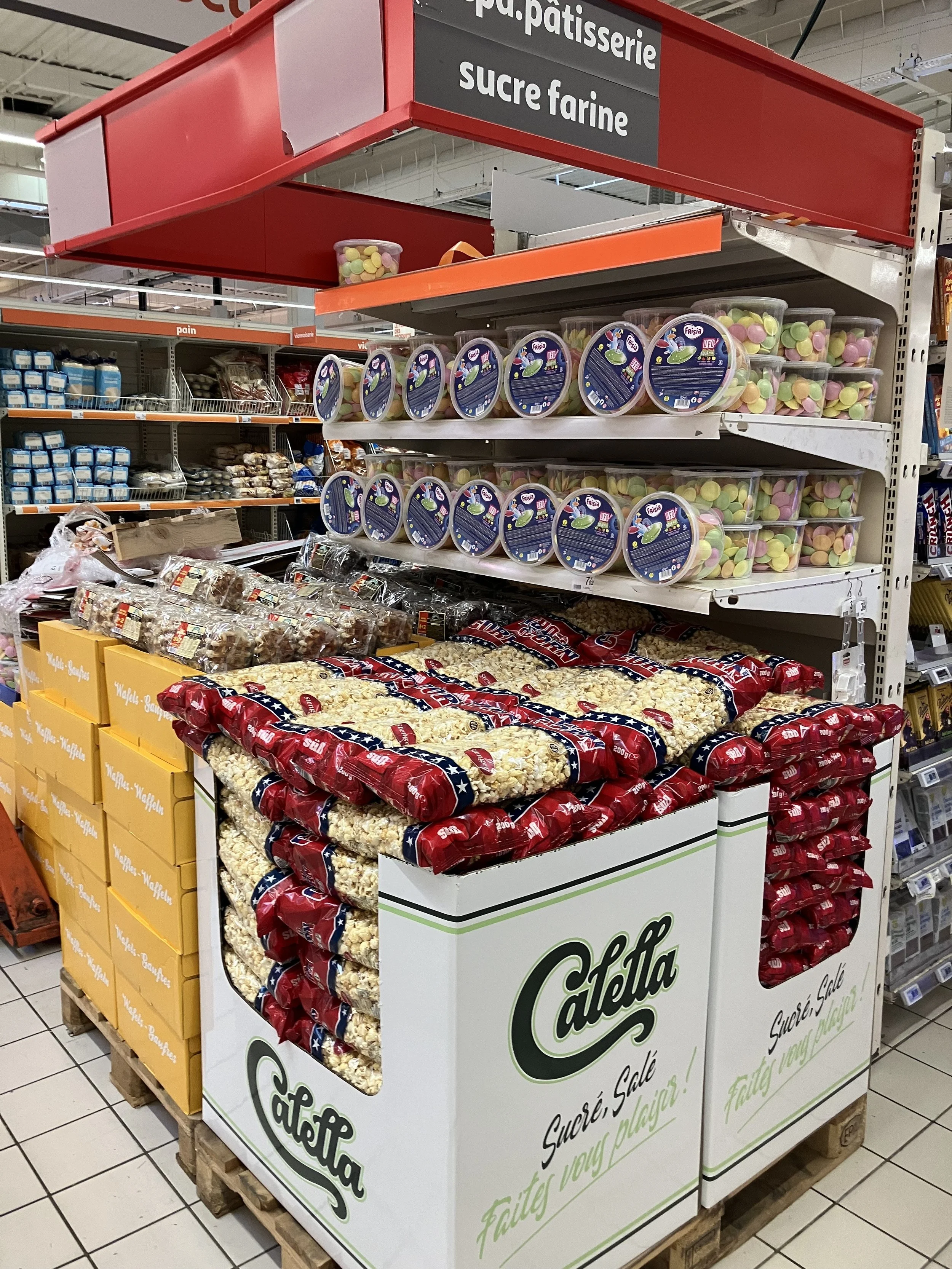 Étagère en supermarché avec des sacs de popcorn, des bonbons en plastique, et des friandises, sous un panneau indiquant 'pâtisserie', 'sucre', et 'farine'.