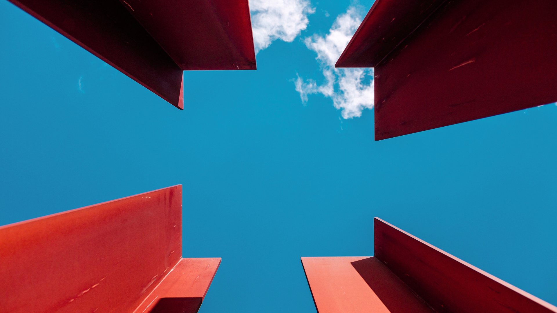 Looking up at a bright blue sky through a square opening framed by four red panels, two on the top and two on the bottom, slightly tilted.