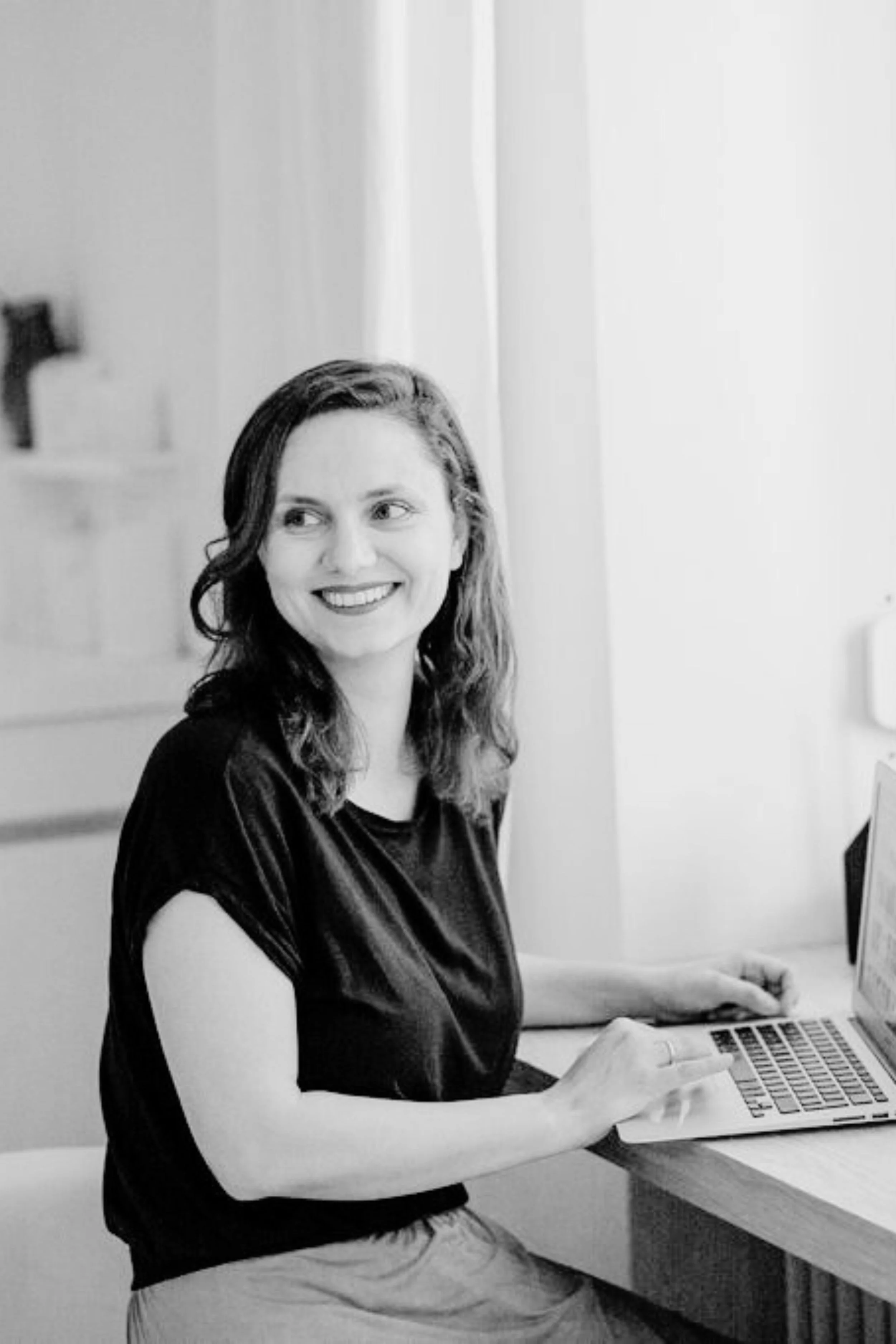 Alexandra Polunin sitzt am Schreibtisch vor einem geöffneten Laptop und dreht sich lachend über die rechte Schulter