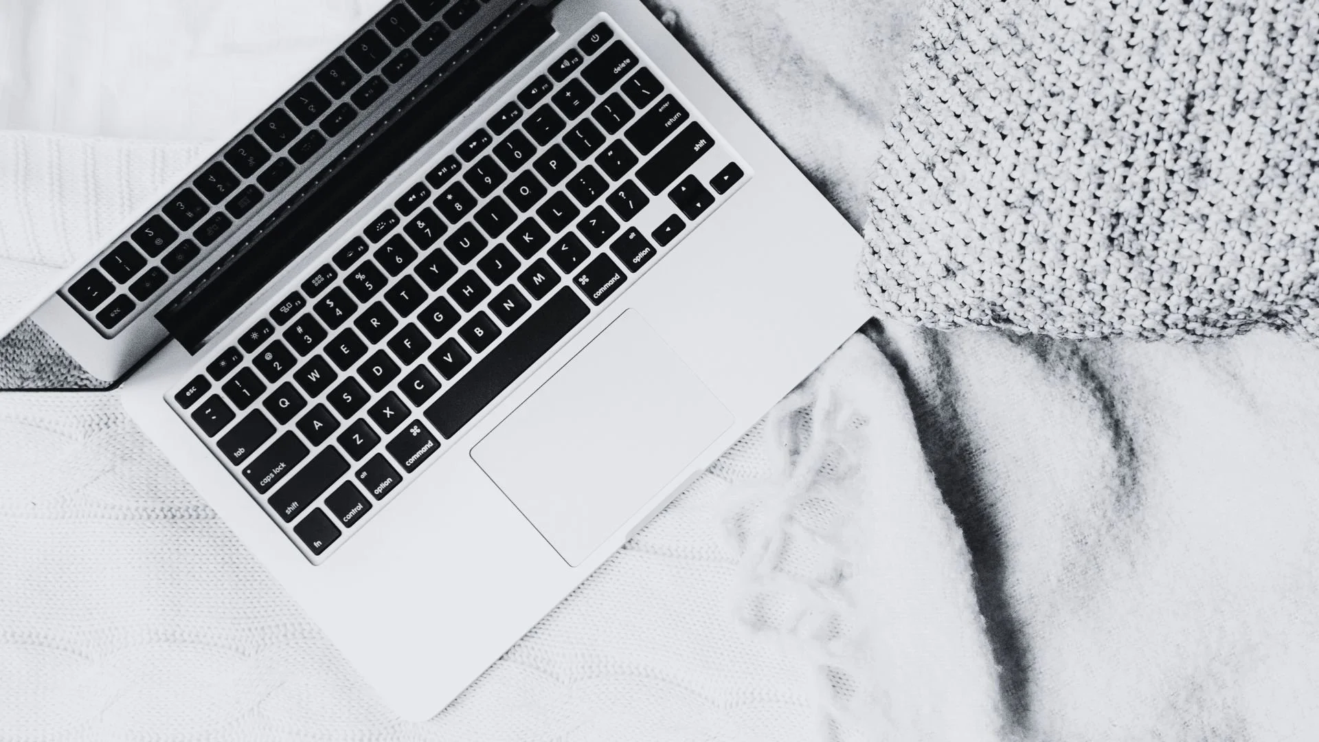 Geöffneter Laptop auf einem weißen Strickdeckchen neben einem grob gestrickten Kissen – gemütlicher Arbeitsplatz im Home-Office.