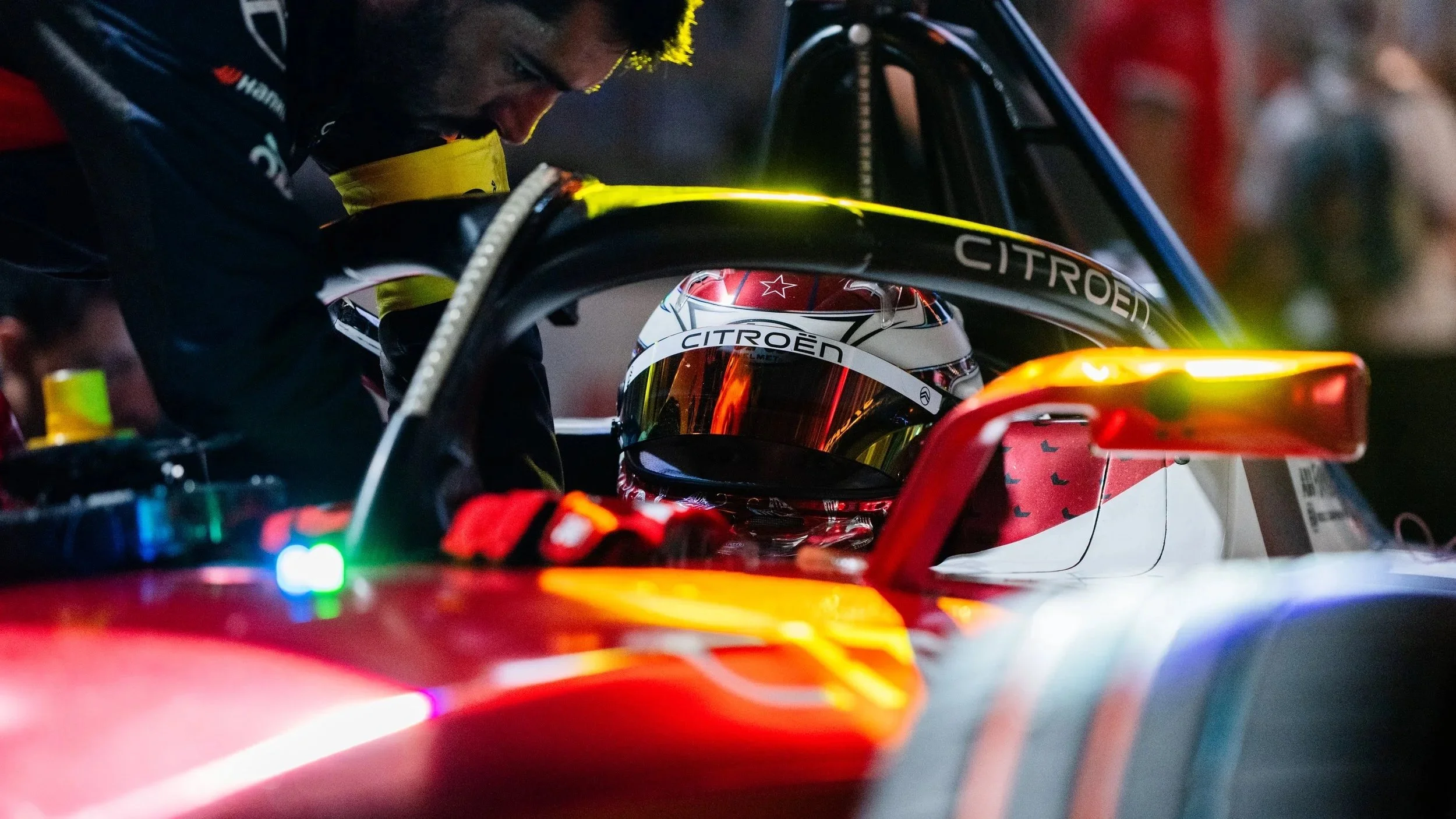 Helmeted Citroën Formula E driver seated in the race car cockpit as a crew member leans over the vehicle.