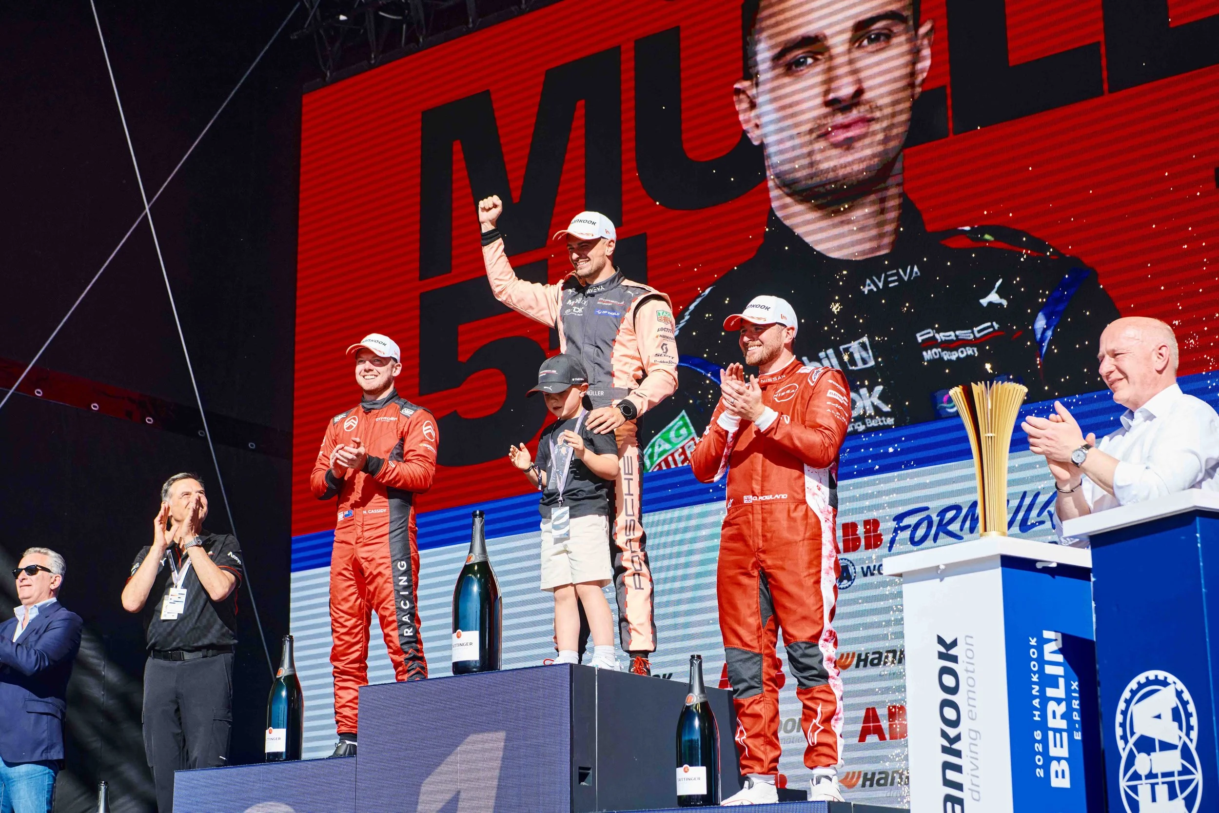 Driver standing on the top step of a podium during a Formula E awards ceremony while other drivers and guests applaud on stage.