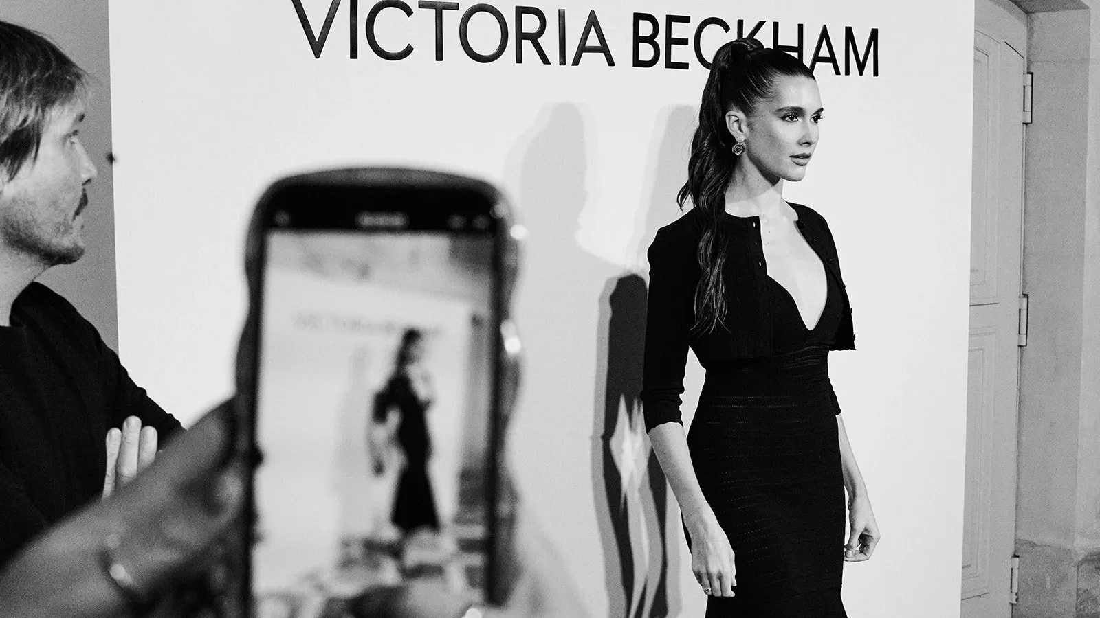 Black-and-white photo of a woman in a black dress posing against a Victoria Beckham backdrop, with a phone capturing her at the Don Julio collaboration event during Paris Fashion Week.