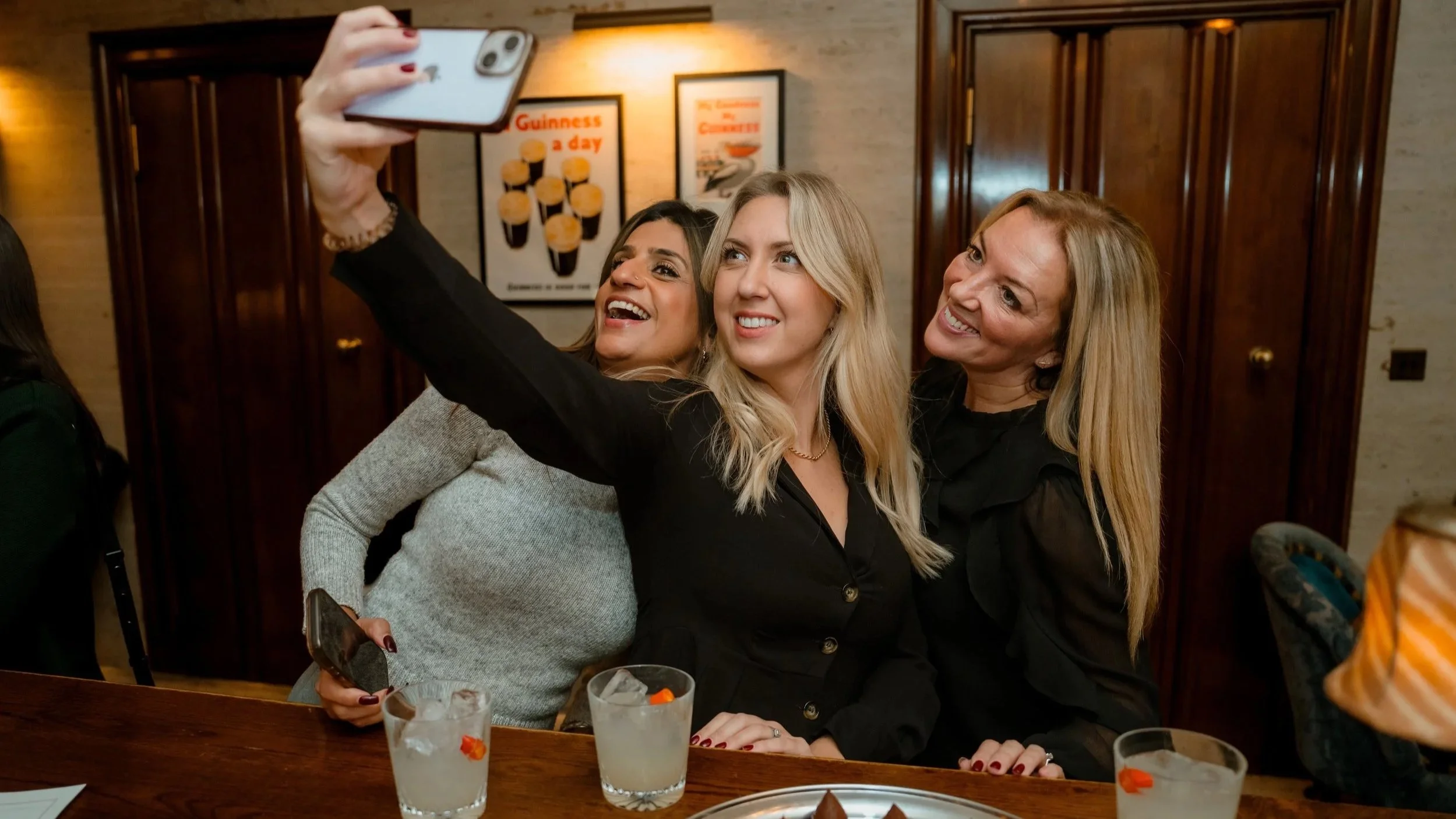 Three women smiling for a selfie beside a bar at the MRA Search corporate networking event.
