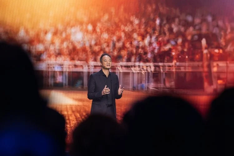 Speaker presenting on stage at a large conference with audience members visible in the foreground.