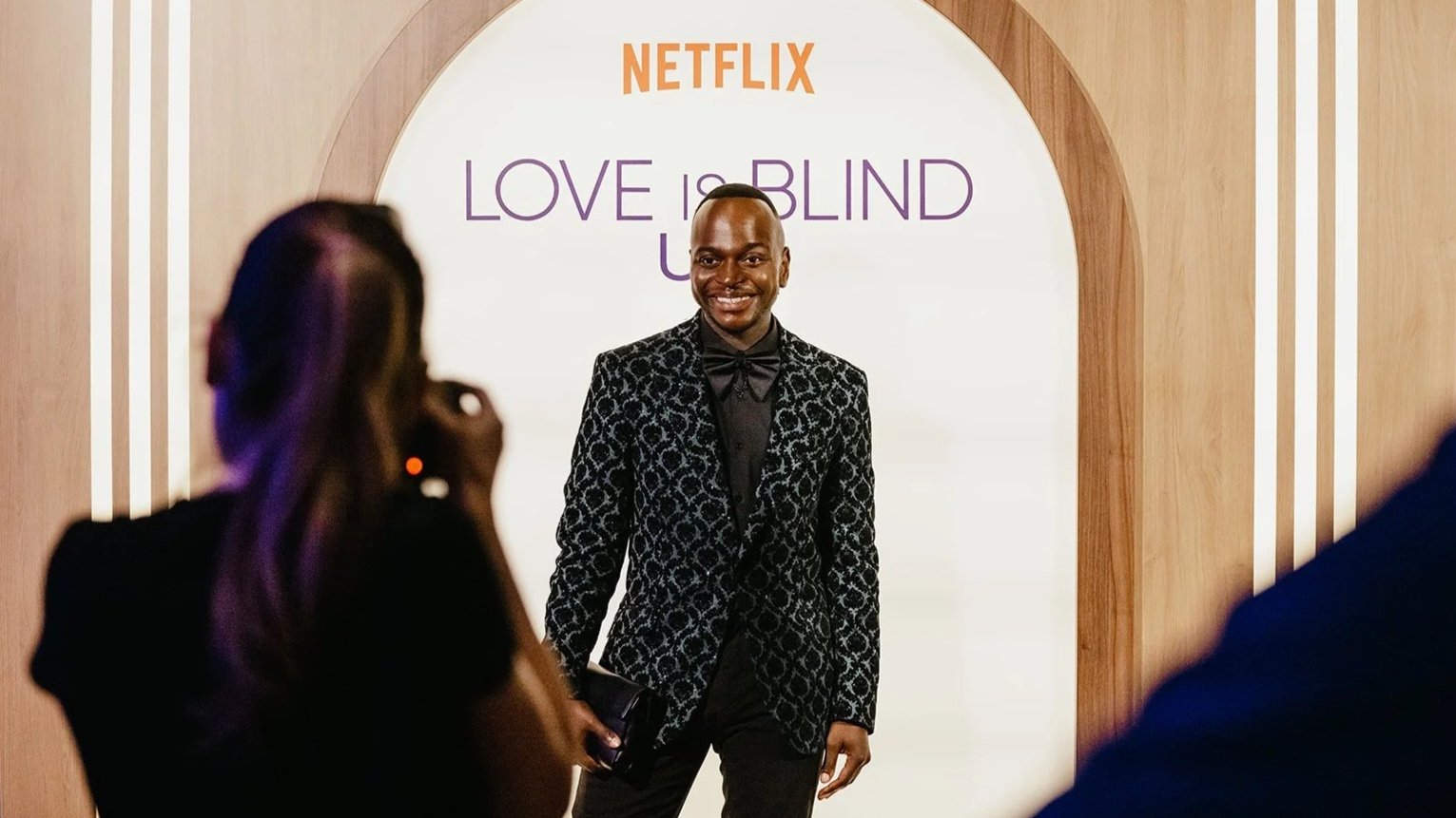 Man in a patterned black jacket posing for a photographer in front of a Netflix Love Is Blind UK backdrop at the London event.